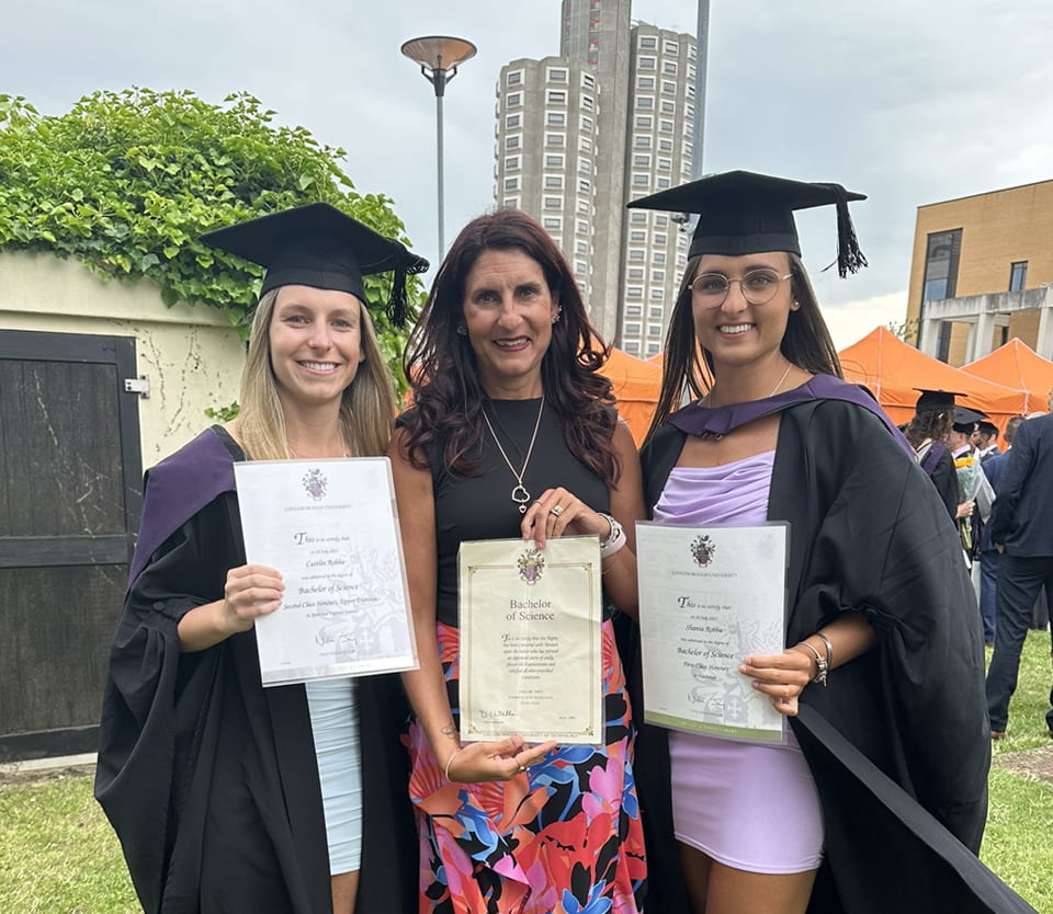 Caitlin, Caroline and Shania Robba each holding their degree certificates. Caitlin and Shania are wearing graduation caps and gowns. The trio stand on grass and in the background is Towers Hall.